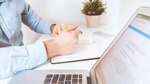 Service (Symbolbild) Zu sehen sind auf einem Schreibtisch liegende Unterarme einer Person, ein Heft und ein Laptop.