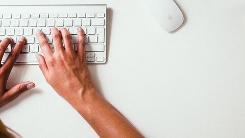 Blick von oben: Auf einer weißen Tastatur, die auf einer weißen Fläche steht, tippt eine Frau.