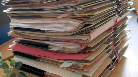 Das Bild zeigt nachlässig aufeinandergestapelte Akten auf einem Holztisch.