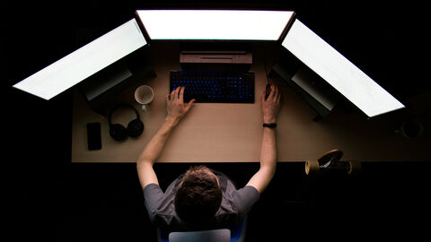 Mann sitzt in einem dunklen Raum vor drei Monitoren.