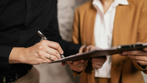 Eine Person unterschreibt ein Schriftstück, das die zweite ihm hinhält.